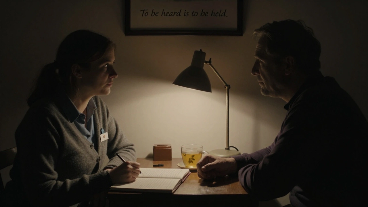 A woman in a cardigan and man sit together in a dimly lit room, sharing a moment of emotional connection.